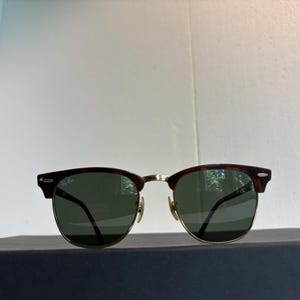 May include: Clubmaster sunglasses featuring a dark tortoiseshell browline and gold-tone metal rims. The lenses are dark green, and the Ray-Ban logo is printed in white on the upper right lens. The frame includes gold-tone nose pads and metal hinges.