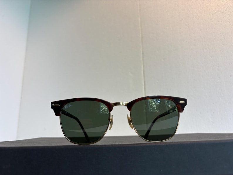 May include: Clubmaster sunglasses featuring a dark tortoiseshell browline and gold-tone metal rims. The lenses are dark green, and the Ray-Ban logo is printed in white on the upper right lens. The frame includes gold-tone nose pads and metal hinges.