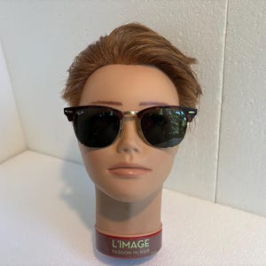 May include: Clubmaster-style sunglasses featuring dark tortoiseshell browline frames, gold-tone metal rims, and dark green lenses. The Ray-Ban logo is visible on the upper corner of the right lens. The glasses are displayed on a mannequin head.