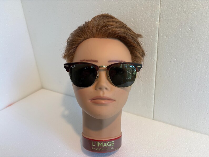 May include: Clubmaster-style sunglasses featuring dark tortoiseshell browline frames, gold-tone metal rims, and dark green lenses. The Ray-Ban logo is visible on the upper corner of the right lens. The glasses are displayed on a mannequin head.