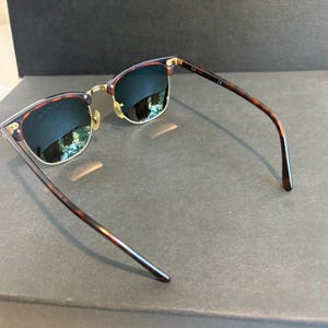 May include: Clubmaster-style sunglasses featuring dark tortoiseshell patterned frames with gold-tone metal accents. The lenses are dark and reflective, and the temples are thin, matching the tortoiseshell pattern of the browline frame.