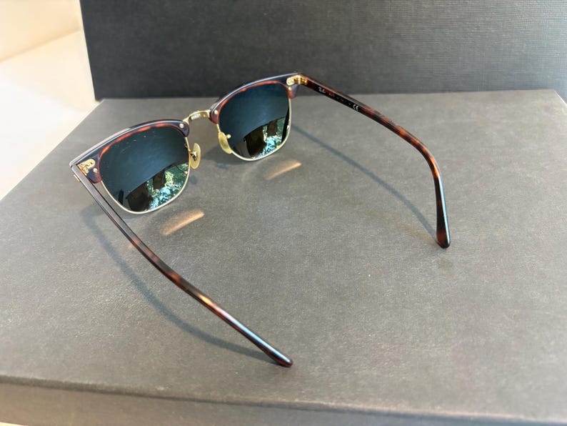 May include: Clubmaster-style sunglasses featuring dark tortoiseshell patterned frames with gold-tone metal accents. The lenses are dark and reflective, and the temples are thin, matching the tortoiseshell pattern of the browline frame.