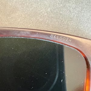 May include: Close-up of sunglasses with a brown tortoiseshell frame. The text "B&L5024" is printed on the frame. The lenses are dark, and the overall design is classic and stylish.