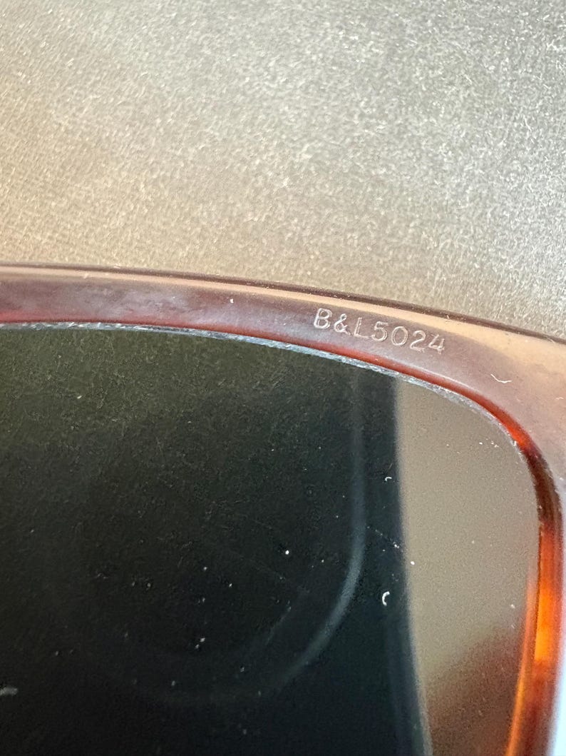 May include: Close-up of sunglasses with a brown tortoiseshell frame. The text "B&L5024" is printed on the frame. The lenses are dark, and the overall design is classic and stylish.