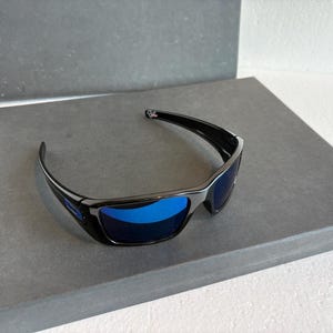 May include: Black sunglasses with blue lenses rest on a gray surface. The sunglasses have a sleek, modern design with a curved frame and a small emblem on the temple. The lenses are a vibrant blue color.