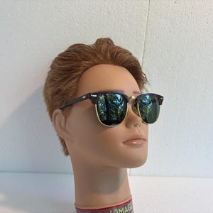 May include: Browline sunglasses featuring dark brown upper frames and gold-tone lower rims. The lenses are dark green and reflective, showing a tree-lined outdoor scene. The glasses are displayed on a mannequin head with short brown hair.