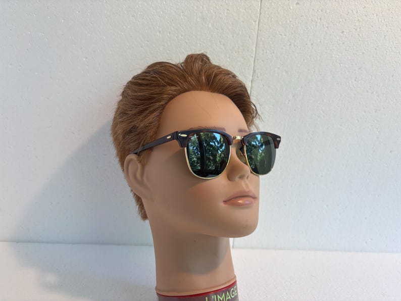 May include: Browline sunglasses featuring dark brown upper frames and gold-tone lower rims. The lenses are dark green and reflective, showing a tree-lined outdoor scene. The glasses are displayed on a mannequin head with short brown hair.