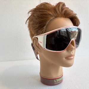 Puede incluir: Gafas de esquí con montura blanca y roja y lentes oscuras. Las gafas se muestran en una cabeza de maniquí con cabello castaño. Las lentes reflejan el entorno. Las gafas tienen una correa con la palabra "CAVA" impresa.
