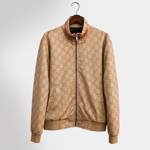 Könnte beinhalten: Beige Reißverschlussjacke mit sich wiederholendem braunen "GG" Logo-Muster. Die Jacke hat einen schwarzen Reißverschluss, einen rot-schwarz gestreiften Kragen und braune gerippte Bündchen und Saum. Die Jacke hängt an einem Metallbügel.