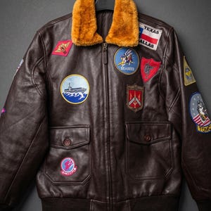 May include: A brown leather bomber jacket with a tan fur collar and various embroidered patches. The jacket has two front pockets and a ribbed hem. Patches include the word "Texas" and military-themed designs.