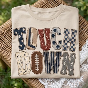 May include: A beige crew neck t-shirt featuring the word TOUCHDOWN in large block letters. Each letter has a unique pattern including stars, plaid, football play diagrams, helmets, checkers, a football, stripes, and lightning bolts.
