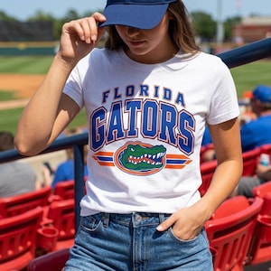 May include: A white short-sleeved t-shirt featuring the text FLORIDA GATORS in blue and orange block lettering, with a graphic of a green alligator head centered below the text, all printed on the front of the cotton fabric.