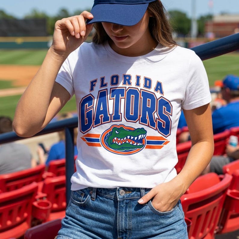 May include: A white short-sleeved t-shirt featuring the text FLORIDA GATORS in blue and orange block lettering, with a graphic of a green alligator head centered below the text, all printed on the front of the cotton fabric.