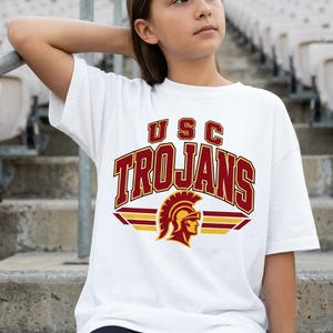 May include: A white short-sleeved t-shirt featuring a graphic print with the text USC TROJANS in dark red with gold outlines. Below the text is a dark red and gold Trojan helmet icon centered above two horizontal gold and dark red stripes.