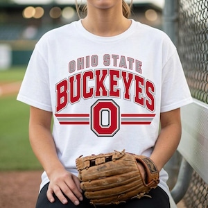 May include: A white short-sleeved t-shirt featuring the text OHIO STATE BUCKEYES in red and grey block lettering, with a large red letter O centered below the text, flanked by two horizontal red and grey stripes on each side of the letter.