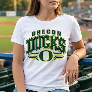 May include: A white short-sleeved t-shirt featuring the text OREGON DUCKS in large green block letters with yellow outlines, positioned above a large green letter O with yellow wing-like accents on a white background.