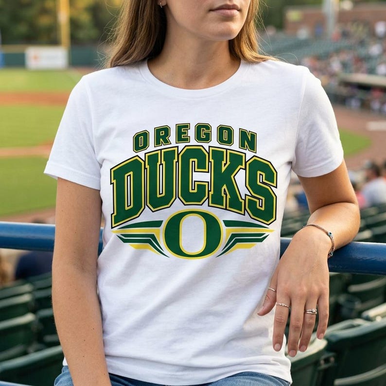 May include: A white short-sleeved t-shirt featuring the text OREGON DUCKS in large green block letters with yellow outlines, positioned above a large green letter O with yellow wing-like accents on a white background.