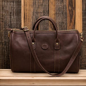 May include: A dark brown leather duffel bag featuring two top handles, a detachable shoulder strap with a gold-toned buckle, a top zipper closure, and a small oval-shaped leather patch centered on the front panel with visible contrast stitching.