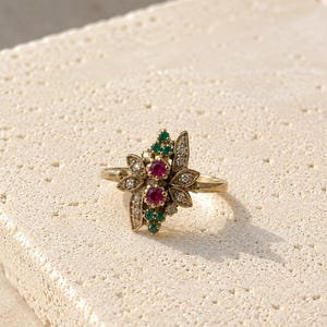 Op de afbeelding: Een goudkleurige ring met een bloemmotief, bezet met robijn, smaragd en diamant. Het middelpunt van de ring toont een cluster van rode robijnen en groene smaragden, omringd door kleine diamanten. De ring heeft een delicate, vintage uitstraling.
