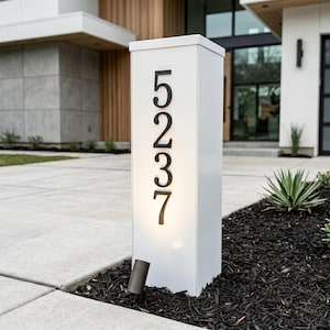 Può includere: Un palo indirizzo rettangolare bianco con il numero "5237" in nero. Il palo ha una luce in basso ed è inserito in un letto di pacciame nero. Lo sfondo include una casa moderna con rivestimento in legno.