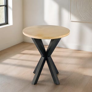 May include: A round table with a light-toned wooden top and a black metal base. The base consists of four angled legs that intersect in the center, creating an X-shaped support structure that provides stability for the circular tabletop surface.