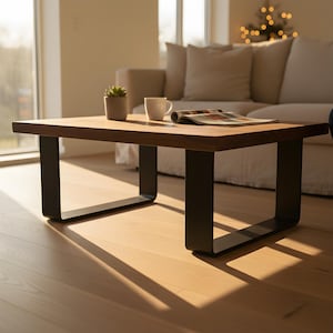 Op de afbeelding: Een rechthoekige salontafel met een donkerhouten blad en zwarte metalen poten. Een kleine plant in een pot, een witte mok en een tijdschrift liggen op de tafel. De tafel staat in een woonkamer met een bank en natuurlijk licht.