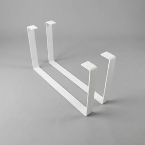 May include: Two U-shaped metal table legs finished in a matte white powder coating. Each leg features a flat mounting plate at the top with pre-drilled holes for attachment, connected to two vertical supports joined by a long horizontal base bar.