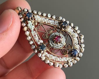 May include: A teardrop-shaped pendant featuring intricate gold filigree metalwork, embellished with numerous small white seed pearls, four dark blue sapphire stones, and a single central red ruby stone set within a circular pearl border.