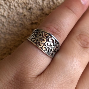 以下が含まれることがあります： 繊細で複雑なハートと渦巻きが絡み合ったデザインが施された、幅広のシルバーリング。バンドは片側がわずかに広くなっており、さりげない非対称性を生み出しています。