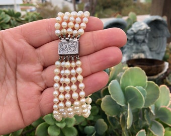 May include: A multi-strand pearl bracelet with a silver clasp featuring an engraved design. The bracelet is made of white pearls and has a classic look.