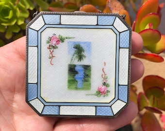 Könnte beinhalten: Ein kleines, quadratisches, silbernes Medaillon mit einem blauen und weißen Emaille-Design. Das Medaillon zeigt eine gemalte Szene mit einer Palme und einem Fluss, mit rosafarbenen Rosen auf beiden Seiten.