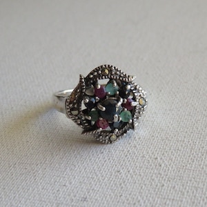 Puede incluir: Un anillo de plata con un grupo de piedras preciosas de colores, incluyendo azul, verde y rojo, engastadas en un delicado diseño de filigrana.