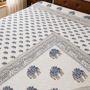 May include: A white quilted bedspread featuring a repeating pattern of small blue and grey elephants. The border displays a larger, more detailed elephant motif in blue and grey, complemented by intricate floral and geometric patterns in blue.