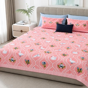 Op de afbeelding: Een roze sprei met een wit golvend latwerkpatroon, witte zwaanillustraties en blauwe bloemmotieven. De set bevat twee bijpassende roze rechthoekige kussenslopen met vergelijkbare patronen, twee lichtblauwe kussens en één klein marineblauw kussen.