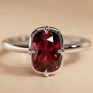 May include: A silver ring featuring a large, oval-cut, deep red gemstone. The gemstone is set in a bezel setting with decorative accents. The band is smooth and polished, reflecting light. The ring is displayed against a neutral background.