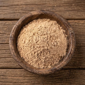 May include: A round, dark brown wooden bowl filled with a mound of fine, light tan powder. The bowl has a visible wood grain texture and a rustic appearance, containing a dry, uniform substance that fills the interior space completely.