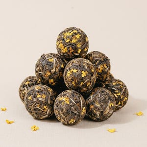 Puede incluir: Una pila piramidal de diez bolas de té redondas compuestas por hojas de té secas de color marrón oscuro entrelazadas con pequeños pétalos de flores de color amarillo brillante. Las bolas de té son texturizadas y esféricas.