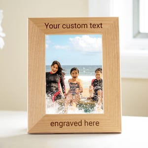 May include: A rectangular wooden picture frame with a light brown finish. The frame holds a photo of three people at the beach. The text "Your custom text" is at the top, and "engraved here" is at the bottom.