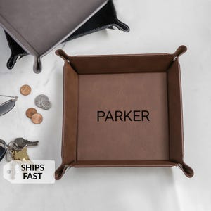 May include: A brown leather valet tray with the name "PARKER" embossed in black. The tray has a square shape with rounded corners and raised edges. Other trays are visible in the background, along with coins, keys, and sunglasses.