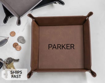 May include: A brown leather valet tray with the name "PARKER" embossed in black. The tray has a square shape with rounded corners and raised edges. Other trays are visible in the background, along with coins, keys, and sunglasses.