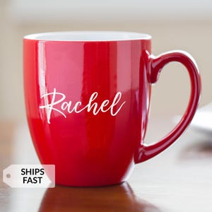 May include: A glossy red ceramic mug with a white interior and a curved handle. The name "Rachel" is written in cursive on the front. A white tag with the words "SHIPS FAST" is in the lower left corner.