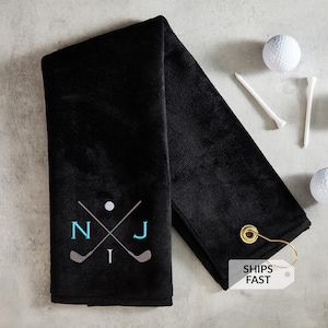 May include: Black golf towel with embroidered golf clubs, a golf ball, and the initials "NJ" in teal. Two golf balls and two golf tees are next to the towel. A gold-colored hook is attached to the towel.