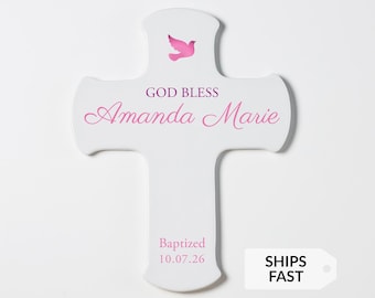 May include: A white cross-shaped wall plaque featuring a pink dove graphic at the top. The text reads GOD BLESS in dark pink, followed by the name Amanda Marie in cursive script, and the words Baptized 10.07.26 in pink at the bottom.