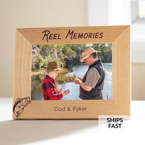 May include: A light-colored wooden picture frame with the words "Reel Memories" engraved at the top. The frame holds a photo of two people fishing. The words "Dad & Ryker" are at the bottom. A fish graphic is on the left.
