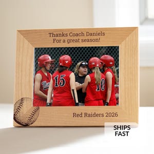 May include: A light-colored wooden picture frame featuring an engraved baseball in the bottom left corner. The top text reads "Thanks Coach Daniels For a great season!" and the bottom text reads "Red Raiders 2026". The frame holds a photo of a softball team.