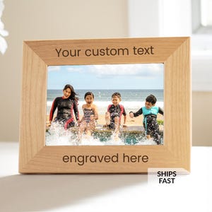 May include: A light-colored wooden picture frame with the text "Your custom text" and "engraved here". The frame holds a photo of four people at the beach. The frame is rectangular.