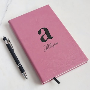 May include: A pink journal with the letter "a" and the name "Allison" printed in black. A black pen with silver accents rests beside the journal. The journal has a textured cover and a black ribbon bookmark.