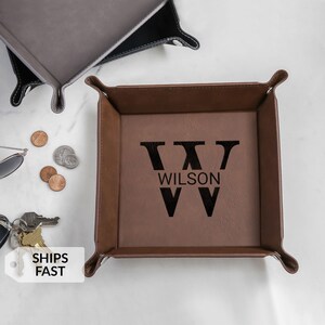 May include: A brown leather valet tray with the name "WILSON" and a large "W" printed in black. The tray has a square shape with rounded corners. Other trays and coins are also visible.
