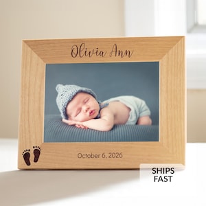 May include: A light-colored wooden picture frame featuring the engraved name Olivia Ann at the top and the date October 6, 2026, at the bottom. The frame includes a small graphic of two baby footprints and holds a photograph of a sleeping infant.