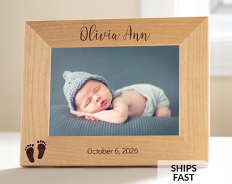 May include: A light-colored wooden picture frame featuring the engraved name Olivia Ann at the top and the date October 6, 2026, at the bottom. The frame includes a small graphic of two baby footprints and holds a photograph of a sleeping infant.